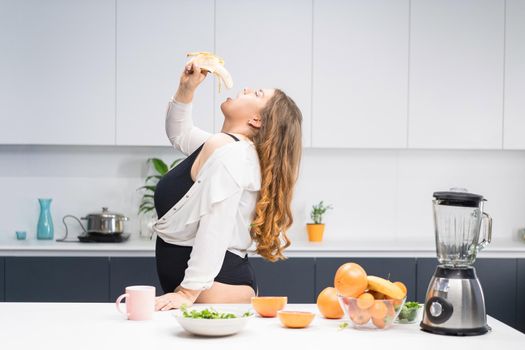 Young fat girl eating banana. Girl sitting on the kitchen table. Diet young fat woman standing on kitchen. Close up chubby female lowering BMI weight. Trying to loose weight fast healthy food.