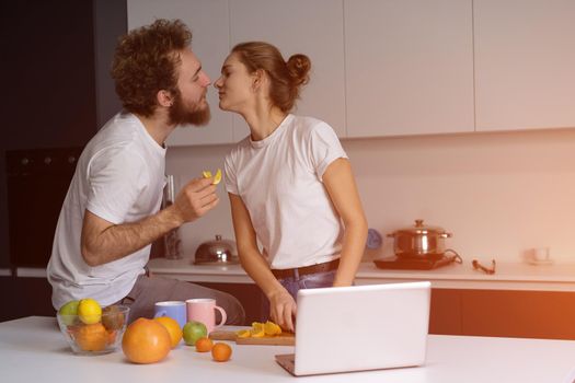 Beautiful young couple kissing feeding each other making fun at modern kitchen and smiling while cooking at home. Pretty girl feeds or nursing her boyfriend.
