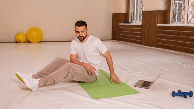 Young handsome man doing crunches and looking online work out videos on using laptop. Self motivated young man warming up doing special exercises for abs muscles.