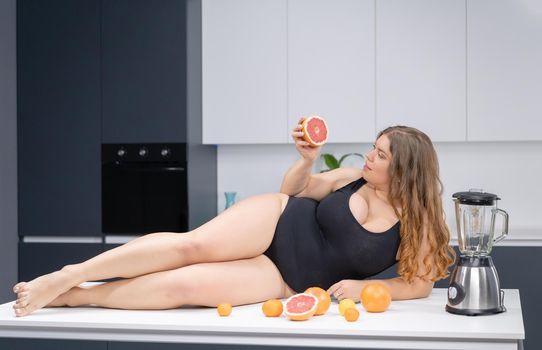 Sexy overweight girl laying on the kitchen table. An obese young sexy chubby white girl in black swimsuit at modern kitchen with fruits next to her.