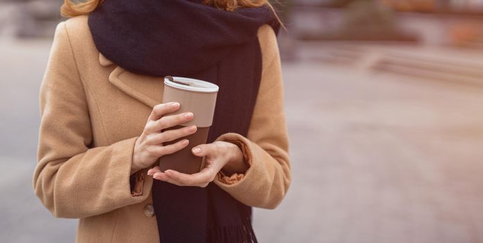 Portrait of a woman's hands holding a coffee mug wearing beige coat and black scarf standing outdoors. Stylish young woman wearing autumn outfit with urban city background. Tinted image.