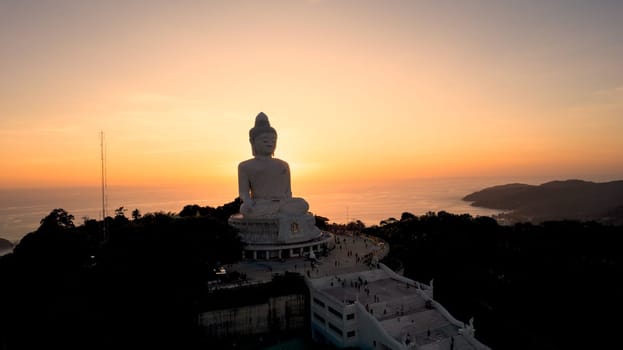 Big Buddha at sunset view from a drone. Phuket. People climb the steps to the statue. The green hills of the island are all around. In the distance, the bright sun goes over the sea. View from above