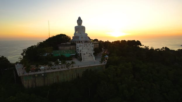 Big Buddha at sunset view from a drone. Phuket. People climb the steps to the statue. The green hills of the island are all around. In the distance, the bright sun goes over the sea. View from above