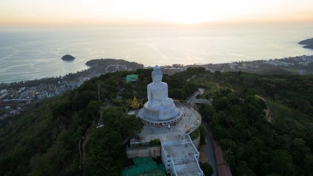 Big Buddha at sunset view from a drone. Phuket. People climb the steps to the statue. The green hills of the island are all around. In the distance, the bright sun goes over the sea. View from above