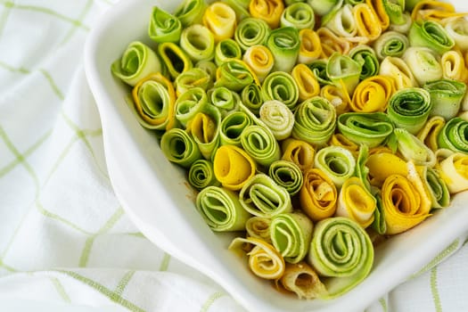 Small rolls of zucchini green and yellow with creamy sauce in a white baking dish against the background of a checkered napkin
