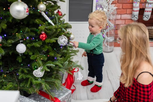 Blurred foreground. In the foreground a woman with her back turned. The woman is kneeling by a Christmas tree. Next to her stands a little boy. Both are hanging Christmas ornaments on the tree. There are gifts lying under the Christmas tree.