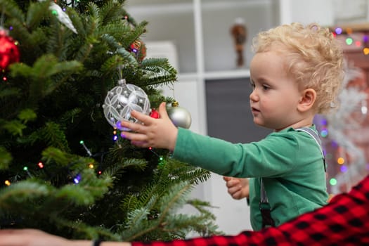 A young child stands by a Christmas tree and extends his hand toward a bauble hanging on a branch. in the foreground you can see a fragment of another person's sleeve. In the background is a bookcase with white shelves and Christmas decorations.