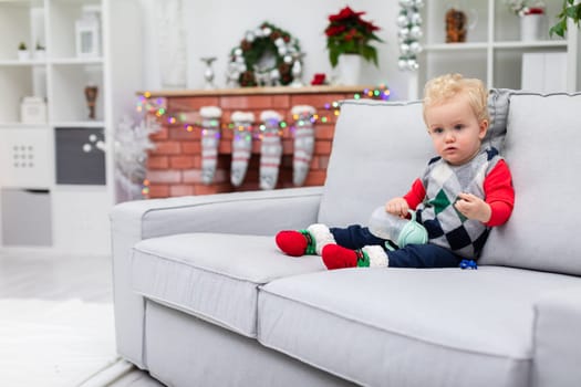 The boy is sitting on a gray couch. In the background you can see a brick fireplace with Christmas decorations hanging on it: colorful lights and Christmas socks. The child is wearing a colorful Christmas sweater and holding a baby bottle in his hand.