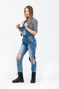 A young girl poses for a photo in a standing position., The girl is wearing a checked shirt and rubbed jeans. The girl is wearing black high top shoes. She is styled in a braid and has disheveled hair.