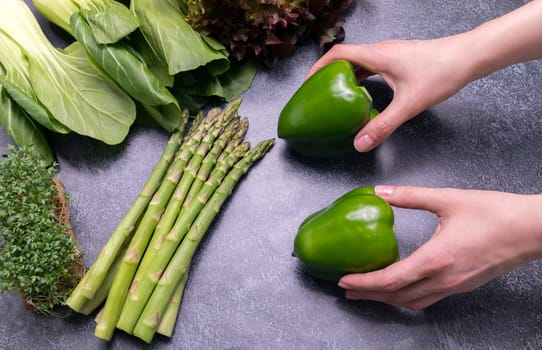 Female Hands Hold Assortment Of Fresh Organic Home Grown Green Vegetables On Granite Table. Asparagus Plant, Bell Pepper, Bok Choy, Red Leaf Lettuce, Bittercresses. Bio food. Horizontal plane