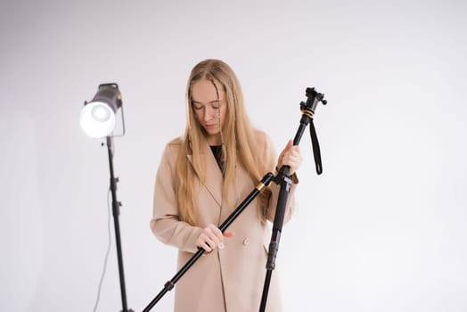 A woman producer, videographer, gaffer a blonde with a tripod and light in the photo studio. Wearing a formal nude pantsuit on a white background