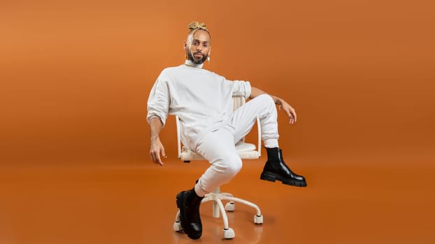 Trendy black latin gay man in white clothes sitting chair look camera isolated on orange background studio portrait People lifestyle fashion lgbtq concept