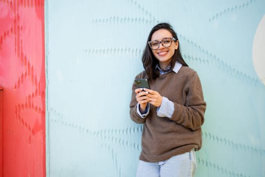 Satisfied hipster girl wear glasses, types text message on mobile phone, enjoys online communication, types feedback, looking at camera, isolated on blue color background. Technology concept
