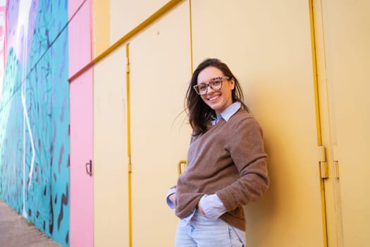 Happy woman in glasses outdoor on yellow color background side view. Positive people concept. Smiling girl looking at camera, hands in pocket, dressed sweater and jeans
