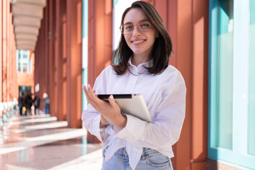 Businesswoman in glasses using digital tablet touching screen looking at camera, standing outdoor dressed white shirt. Female freelancer holding tablet while walk outside