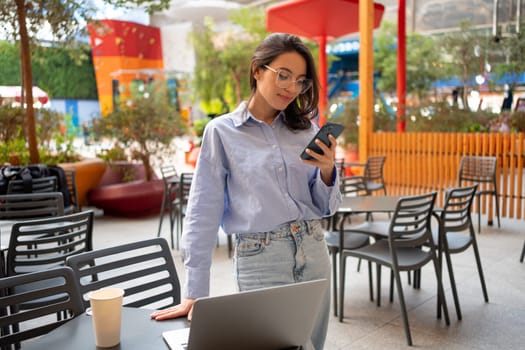Freelance worker doing remote work using smartphone and laptop and smile. Female freelancer with mobile phone standing in outdoor cafe holding phone in hand watching device screen