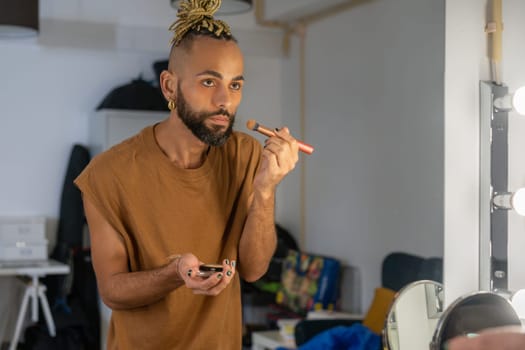 Handsome bearded black gay applying make up on face looking mirror. Transgender non binary person use makeup brush.