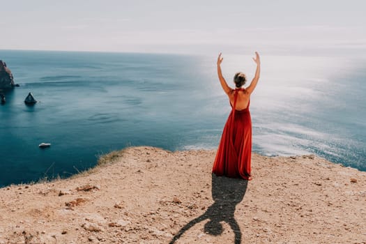 Side view a Young beautiful sensual woman in a red long dress posing on a rock high above the sea during sunrise. Girl on the nature on blue sky background. Fashion photo.