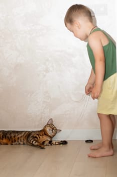 A little beautiful boy of 4 years old is having fun playing with a red, leopard bengal cat on the floor in a home interior. The domestic cat lies on the floor and plays with the baby with its paw.