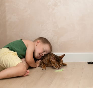 A little beautiful boy of 4 years old is having fun playing with a red, leopard bengal cat on the floor in a home interior. The domestic cat lies on the floor and plays with the baby with its paw.