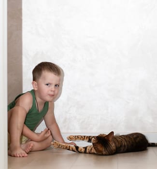 A little beautiful boy of 4 years old is having fun playing with a red, leopard bengal cat on the floor in a home interior. The domestic cat lies on the floor and plays with the baby with its paw.