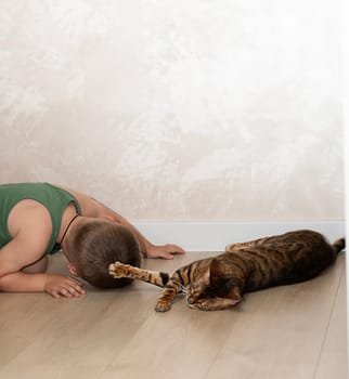 A little beautiful boy of 4 years old is having fun playing with a red, leopard bengal cat on the floor in a home interior. The domestic cat lies on the floor and plays with the baby with its paw.