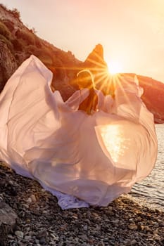 Mysterious woman silhouette long hair walks on the beach ocean water, sea nymph wind listens to the wave. Throws up a long white dress, a divine sunset. Artistic photo from the back without a face.