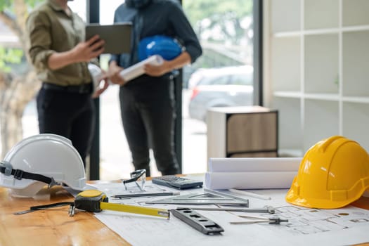 Concept of engineering equipments of protective helmet for competent engineer on piles of paper works. Blurred image of engineer working together at the desk.