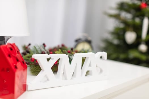 Blurred background. In the background a decorated Christmas tree, a white curtain in the window. In the foreground a Christmas decoration in the form of a white space inscription. Next to the inscription stands a bedside lamp and a red candlestick.