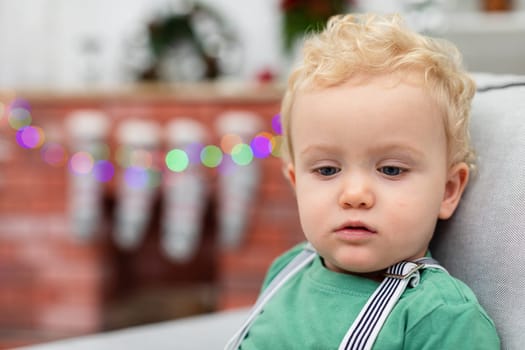 Close-up on the face of a little boy looking down. The boy has curly blond hair and a sleepy face. In the background you can see the blurred outline of a brick fireplace. Christmas ornaments and lights are attached to the fireplace.