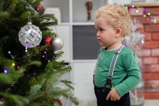 Blurred foreground. In the foreground a woman with her back turned. The woman is kneeling by a Christmas tree. Next to her stands a little boy. Both are hanging Christmas ornaments on the tree. There are gifts lying under the Christmas tree.