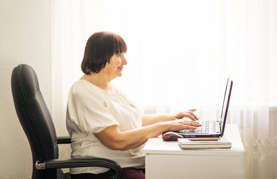 Senior smiling business woman working at her computer laptop at home. Business, education and technology concept. Portrait of a skilled female programmer freelancer with laptop sitting on the table in an office.