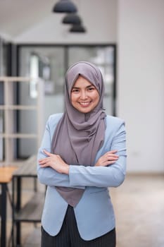 A portrait of a beautiful and professional Asian Muslim businesswoman stands in the office with her arms crossed...