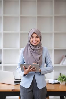 A portrait of a successful Asian Muslim businesswoman stands in her modern office with a digital tablet in her hand...