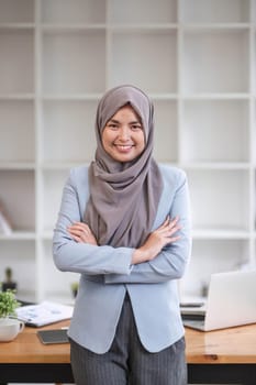 A portrait of a beautiful and professional Asian Muslim businesswoman stands in the office with her arms crossed...
