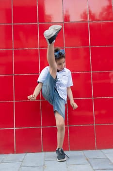 A cute boy in a white shirt raises his leg against a red wall, imitates the game of football, the child actively enjoys an active life, High quality photo