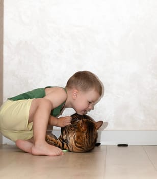 A little beautiful boy of 4 years old is having fun playing with a red, leopard bengal cat on the floor in a home interior. The domestic cat lies on the floor and plays with the baby with its paw.