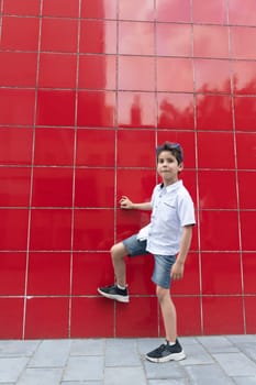 A cute boy in a white shirt raises his leg against a red wall, imitates the game of football, the child actively enjoys an active life, High quality photo