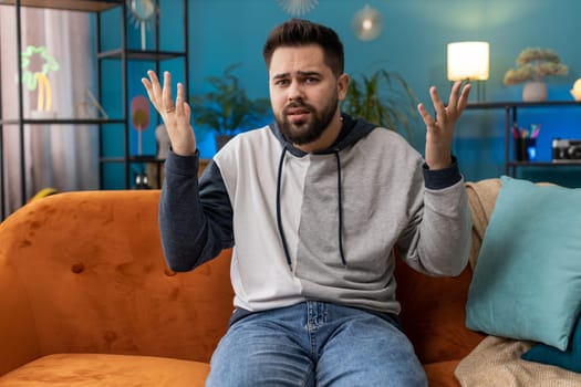 What. Why. Caucasian man raising hands in indignant expression asking reason of failure demonstrating disbelief irritation by troubles at home apartment. Young guy in living room sitting on sofa