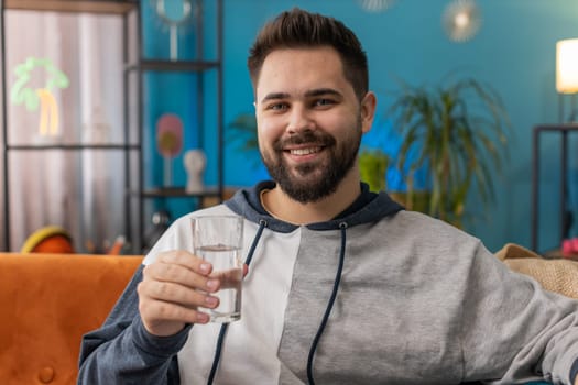 Thirsty man holding glass of natural aqua make sips drinking still water preventing dehydration sits at home living room. Caucasian guy with good life habits, healthy slimming, weight loss concept