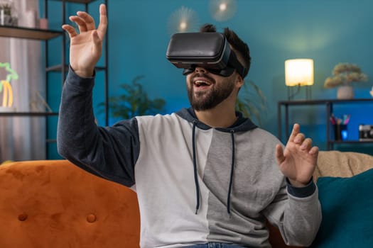 Excited man using virtual reality futuristic technology VR app headset helmet to play simulation 3D 360 video game watching film movie. Portrait of guy in goggles at modern home apartment room on sofa