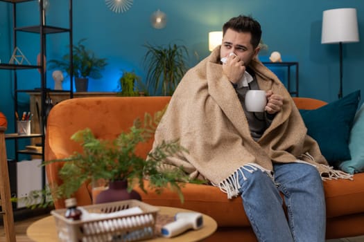 Sick man wrapped in plaid sit alone shivering from cold on sofa drinking hot tea in unheated apartment without heating due debt. Unhealthy ill guy feeling discomfort try to warming up at home room