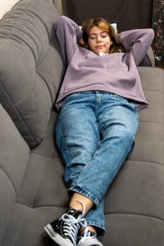 a teenage girl is lying on the couch with her hands behind her head. a girl in blue jeans and sneakers and a purple hoodie lies with a hood on her head, looks at the camera and smiles. A fashionable concept in casual style.