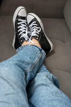 the legs of a teenage girl dressed in blue jeans and new youth sneakers with lacing lie on the home sofa. A fashionable concept in the casual style. vertical photo