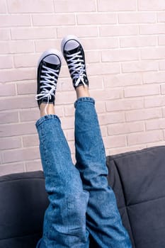 a teenage girl, dressed in blue jeans and new sneakers with lacing, is lying on the sofa with her legs thrown up. A fashionable concept in casual style. rest and relaxation. Mental health.