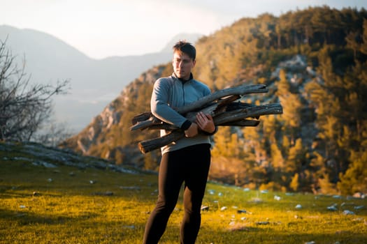 Young attractive man holds firewood, carries it to the camp against the backdrop of a picturesque landscape with hills, mountains and forest at the sunset in the evening. Traveler is hiking outdoor.