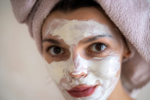 A young girl in a towel on her head and body stands near the mirror in the bathroom and applies a clay mask to her face, the woman takes care of her health and beauty.