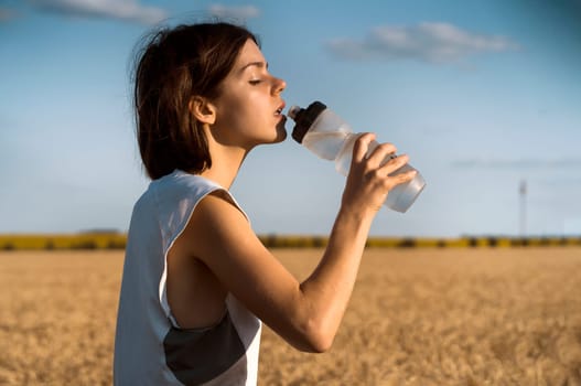 A young girl takes a break during an intense workout and jogging and drinks refreshing cool water from a bottle, a woman trains outdoor in the evening at sunset.