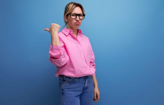 young blond caucasian woman model with a ponytail hairstyle in a stylish look consisting of a pink blouse and jeans frustratedly points her finger to the side.