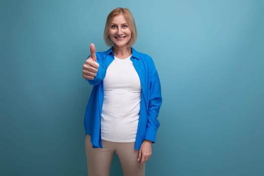 portrait of positive blonde 60s woman in blue stylish shirt showing like on studio background.